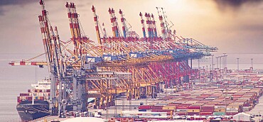 Containerhafen mit vielen Kränen, einem Schiff und unzähligen Containern vor einem rosa Abendhimmel