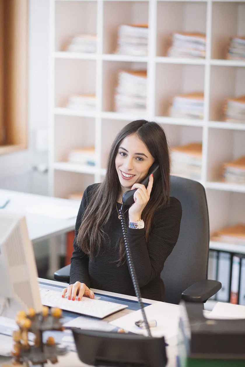 Eine Frau sitzt in einem Büro vor einem Regal mit Akten und Unterlagen und schaut lächelnd in die Kamera, während sie telefoniert.