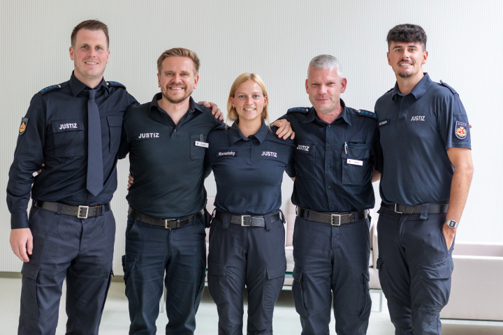 fünf Bedienstete der Justizvollzugsanstalt in Uniform lächeln in die Kamera und haben sich die Arme um die Schultern gelegt
